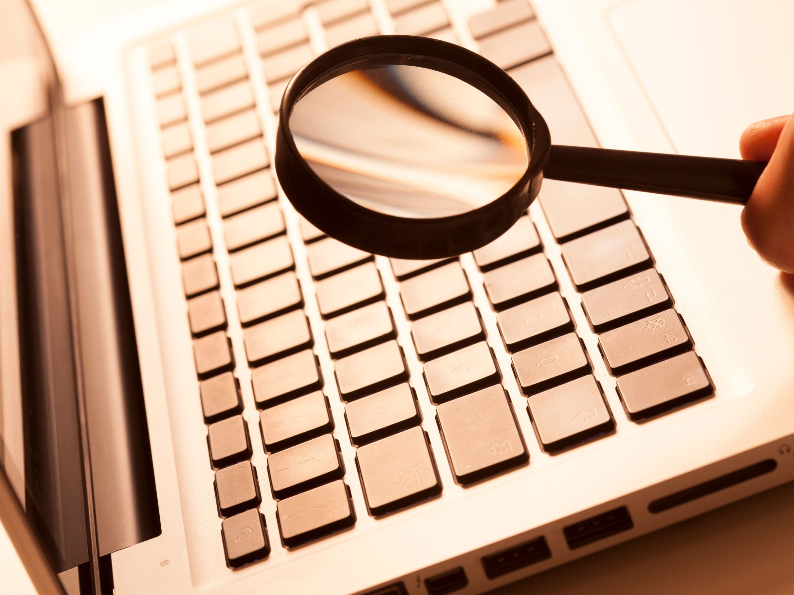 Hand holding a magnifying glass over a laptop keyboard, stock research tools article